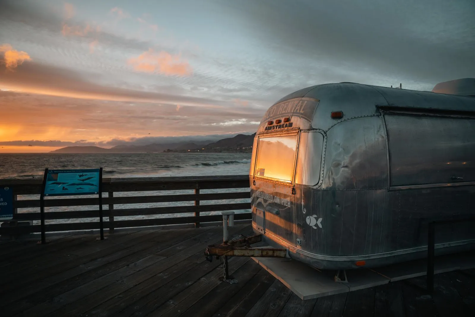 A classic Airstream trailer reflecting a vibrant sunset on an ocean pier.